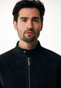 Chaqueta de ante negra con un cuello alto, cremallera frontal y botones a presión. Textura suave, diseño entallado y silueta ajustada.