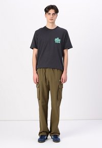 Schwarzes Baumwoll-T-Shirt mit türkisfarbigem Grafikdruck auf der Brust, kombiniert mit olivgrünen Cargo-Hosen, die Taschen und eine lockere Passform aufweisen. Blaue Sneaker.