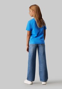 T-shirt di cotone blu a maniche corte abbinata a jeans a gamba larga di un blu chiaro. Sneakers bianche completano il look casual. Visibile da dietro.