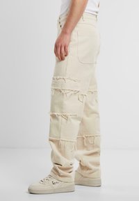 Pantaloni din denim bej, supradimensionați, cu accente destrămate și detalii din petice. Prezintă o croială lejeră și un buzunar pe partea dreaptă.