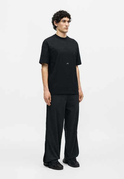 Άντρας που στέκεται στραμμένος προς τα εμπρός, φοράει μαύρο υπερμεγέθες T-shirt, φαρδιά μαύρη παντελόνια και μαύρα αθλητικά παπούτσια σε απλό λευκό φόντο.