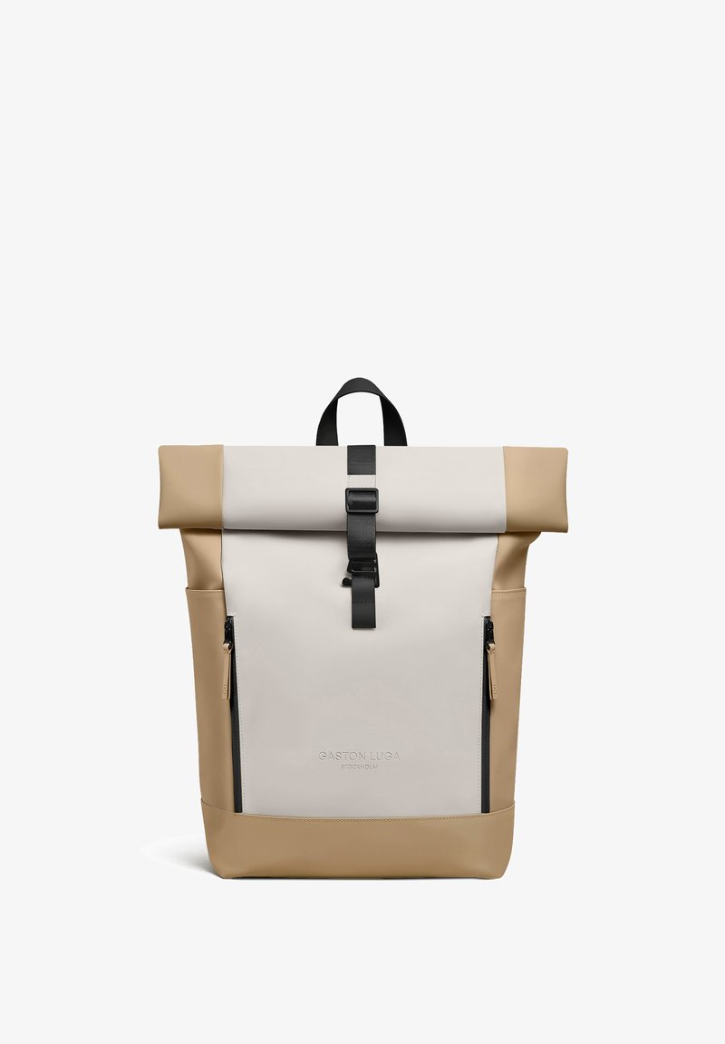 Sac à dos beige et crème à rabat roulant avec boucle noire, deux fermetures éclair verticales à l'avant et poignée, nom de la marque "Gaston Luga Stockholm" embossé à l'avant.