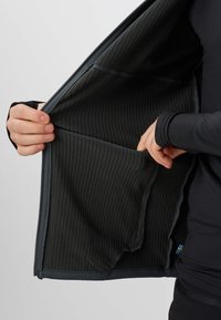 Hænder holder en mørk ribbet lynlåstrøje åben, der viser dens indvendige lomme og en lille svensk flagmærkat nær kanten.