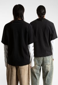 Twee personen dragen zwarte T-shirts met korte mouwen, gelaagd over witte overhemden met lange mouwen, gecombineerd met beige corduroy en lichte denim broeken.
