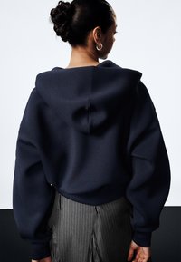 Kvinne med mørkt hår i en knute, iført sølv hoops, en navy oversized hettegenser og et grått stribet skjørt med folder, vendt bort.
