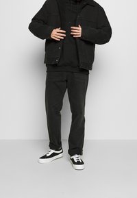 Persona con chaqueta de denim forrada de sherpa negra, sudadera negra, jeans negros y zapatillas blanco y negro, ajustándose la chaqueta con las manos.