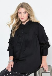 Camicia nera a maniche lunghe con colletto alla cinese e tessuto morbido. Il modello è seduto, indossando pantaloni a pois e gioielli d'argento.