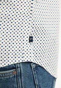 Camicia a maniche corte in tessuto bianco con un motivo geometrico di diamanti blu e verdi; include un piccolo etichetta del marchio sull'orlo.