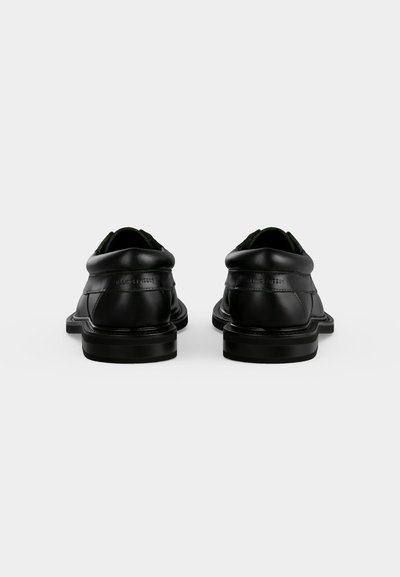 Paire de chaussures habillées en cuir noir vues de dos, avec des cols matelassés et des talons bas empilés sur un fond uni clair.