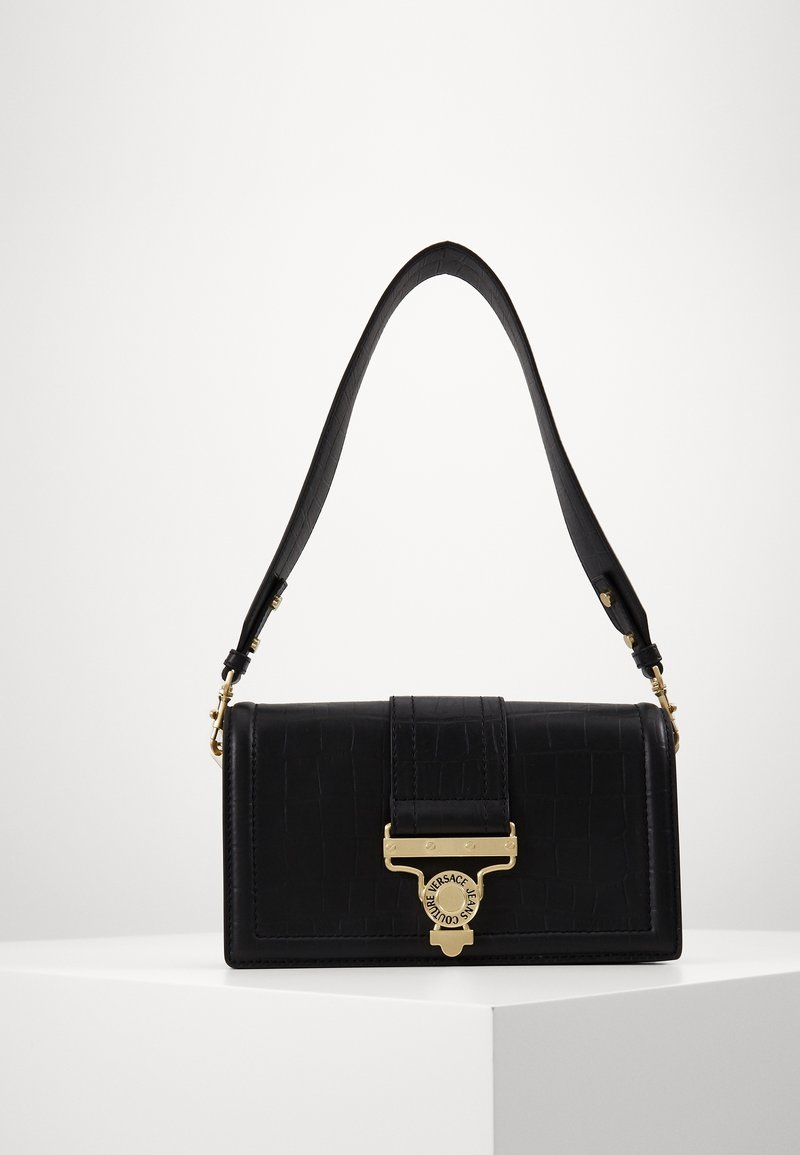 Sac à épaule en cuir noir texturé avec sangle courbée et fermoir doré présentant un logo circulaire sur une surface d'exposition blanche.