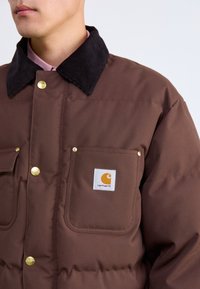 Chaqueta aislante marrón con collar de pana, botones a presión dorados, dos bolsillos frontales y una parche con el logo en el bolsillo izquierdo.