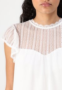 Žena v bílém topu s průhledným krajkovým výstřihem a volnými rukávy, zobrazující detailní květinový krajkový vzor a vroubkovaný výstřih.