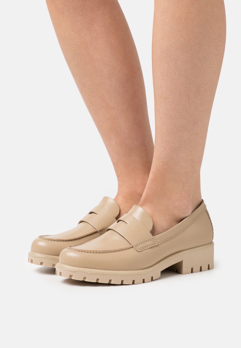 Mocassins en cuir beige avec un bout arrondi, une couture minimale et une semelle texturée, dotés d'un petit talon bloc pour la stabilité.
