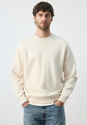 Man met krullend haar, gekleed in een crèmekleurige trui met ronde hals en een blauwe spijkerbroek, staand met handen in de zakken tegen een effen achtergrond.