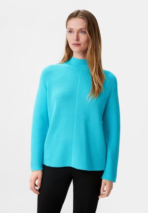 Turkusowy sweter z prążkowanej tkaniny, z wysokim kołnierzem i długimi rękawami. Miękka tekstura i luźny fason, zestawiony z czarnymi spodniami.