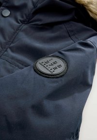 Tissu de veste bleu marine avec une texture lisse, présentant un écusson noir rond sur la manche avec une inscription en relief. Finition en fourrure sur la capuche.