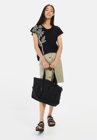 Zwart kortmouwen top met groene bladborduursels, kaki rok met knopen aan de voorkant, zwarte tote bag met ritsen, en zwarte sandalen met banden.