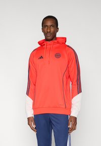 Sweat à capuche de sport rouge avec des rayures bleu marine, logo de la marque sur la poitrine, capuche ajustable et accents blancs sur les manches. Tissu doux et léger.