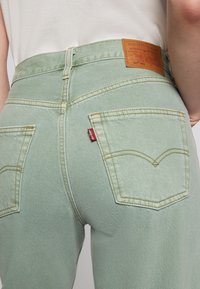 Jean en denim vert avec des coutures jaunes, présentant une étiquette de logo au niveau de la taille et deux poches arrières avec des coutures distinctes.