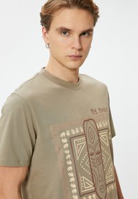 Olivengrøn bomulds T-shirt med en frontgrafik, der viser geometriske mønstre, hænder og en central figur, rund halsudskæring og korte ærmer.