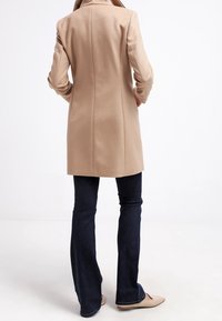 Beige jas met een getailleerde pasvorm, hoge kraag en verticale naden, gecombineerd met donkerblauwe flare jeans en lichtgekleurde ballerina's.