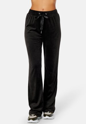 Persona con pantalones de terciopelo negro holgados con cordón de cinta negra y zapatillas deportivas, mostrada desde la mitad del torso hasta los pies sobre un fondo liso.