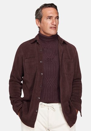 Man wearing dark brown corduroy shirt over a matching cable-knit turtleneck sweater and light-colored pants, hands in pockets, looking to the side.