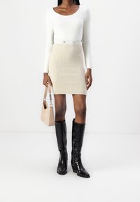 Femme portant un haut blanc à manches longues, une mini-jupe beige texturée avec des boutons, tenant un sac à main beige et des bottes noires montantes avec des talons.