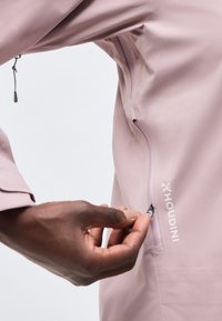 Χέρι με σκουρόχρωμο δέρμα τραβάει το φερμουάρ της τσέπης ενός μωβ-απαλού μπουφάν Houdini, δείχνοντας την υφή του υφάσματος και το λογότυπο της μάρκας στο μανίκι.