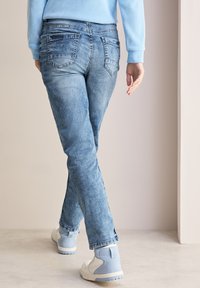 Přiléhavé modré džíny z denimového materiálu s vybledlým vzhledem, pěti kapsami a kontrastním šitím. Nosené s světle modrými teniskami na neutrálním pozadí.