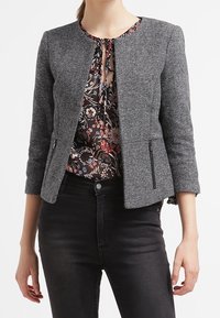 Blazer grigio a motivo houndstooth con colletto rotondo, zip frontali e maniche a tre quarti, indossato sopra una blusa con motivo floreale.