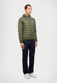 Olivengrønn jakke med hette, quiltet design, frontglidelås og subtil logo. Kombinert med mørk denimjeans og hvite sneakers.
