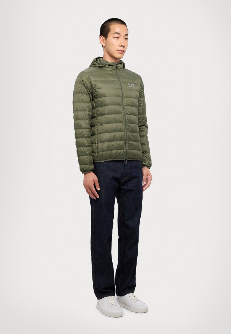 Olivengrønn jakke med hette, quiltet design, frontglidelås og subtil logo. Kombinert med mørk denimjeans og hvite sneakers.