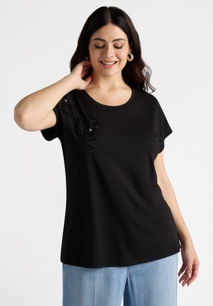 Zwarte korte mouwen t-shirt van een gladde stof, met pailletten bloemenversieringen op de linkerschouder en een ronde halslijn.