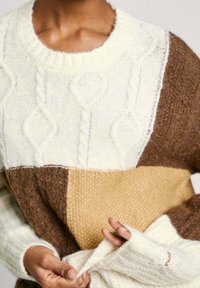 Pull en maille épaisse avec des sections crème, marron et moutarde. Motifs torsadés sur la partie crème et une texture douce partout.