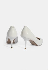 Weiße High Heels mit einer glatten Lederoberfläche, spitzem Zehenbereich und einem schlanken Absatz. Der Absatz hat am Boden eine goldene Akzentuierung.