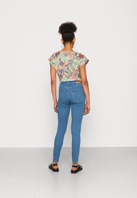Mujer con el cabello rizado en un moño, vistiendo una blusa corta de flores, jeans ajustados azules y sandalias negras, de pie frente a una pared blanca.