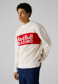 Cremefarbener Pullover mit rotem Band, das "RedBull RACING" in weißem Text zeigt. Gerippte Bündchen und Saum, weiche Textur und lässige Passform.