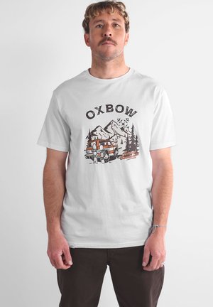 Homme avec une moustache portant un T-shirt blanc avec une montagne, des arbres, un véhicule orange, et l'inscription "OXBOW Beyond Boundaries", se tenant devant un fond uni.