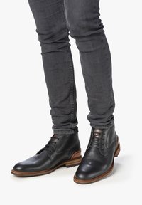 Stivaletti in pelle nera con una texture liscia, punta rotonda e suola in gomma marrone. Presentano lacci e dettagli di design minimalisti.