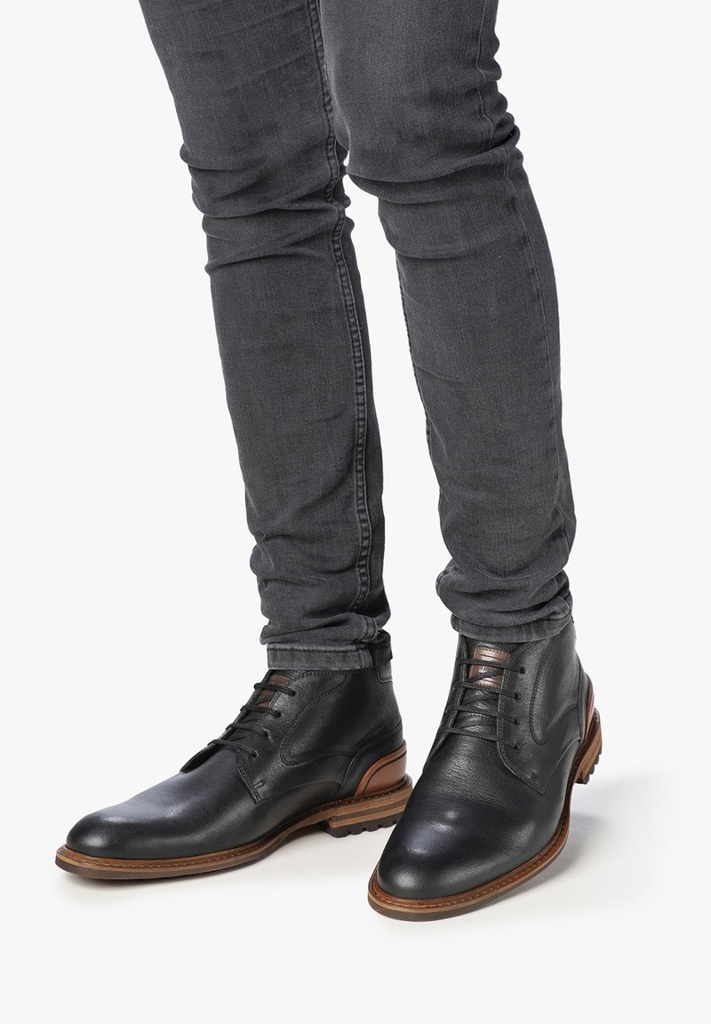 Stivaletti in pelle nera con una texture liscia, punta rotonda e suola in gomma marrone. Presentano lacci e dettagli di design minimalisti.