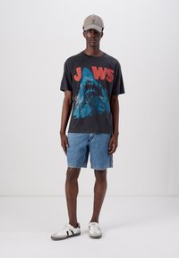 Grafisk JAWS t-skjorte i svart og blå, kombinert med lyseblå jeansshorts og hvite joggesko med grå detaljer. Inkluderer en baseballcap.