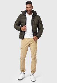 Dunkelgrüne Pufferjacke mit Kapuze und gestepptem Design. Kombiniert mit beigen Jogginghosen und weißen Sneakers, die einen sportlichen Stil zeigen.
