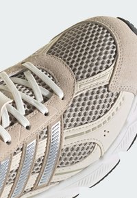 Gros plan d'une chaussure de sport beige et argent avec tissu en mesh, empiècements en daim et lacets blancs sur une semelle blanche avec une bande de roulement noire.