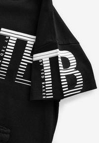 Sort bomulds-T-shirt med hvid grafisk tekst og stribede mønstre på ærmerne, med tydeligt fremhævede mærkeoplysninger.