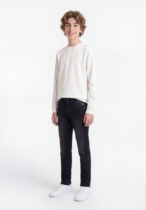 Teenager-Junge steht und lächelt, trägt ein weißes langärmeliges Hemd, schwarze Jeans und weiße Sneakers auf weißem Hintergrund.