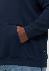 Marineblauwe sweatshirt met een voorzak, ribgebreide manchetten en onderkant. Zachte textuur, met een subtiele merkteken aan de zijkant.