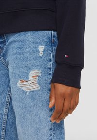 Marinblå sweatshirt med ribbade manschetter som bärs över ljusblå slitna jeans med fransade lappar. Litet logotypdetalj på manschetten.