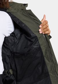 Grüne Außenschaljacke mit schwarzem strukturiertem Innenfutter, ausgestattet mit einer Netzinnentasche und einem Reißverschluss. Glattes Material mit geometrischem Muster.