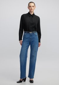 Schwarzes langärmliges Hemd mit Kragen, kombiniert mit hochtaillierten blauen Jeans und schwarzen spitz zulaufenden Absätzen. Goldener Gürtel als Akzent.
